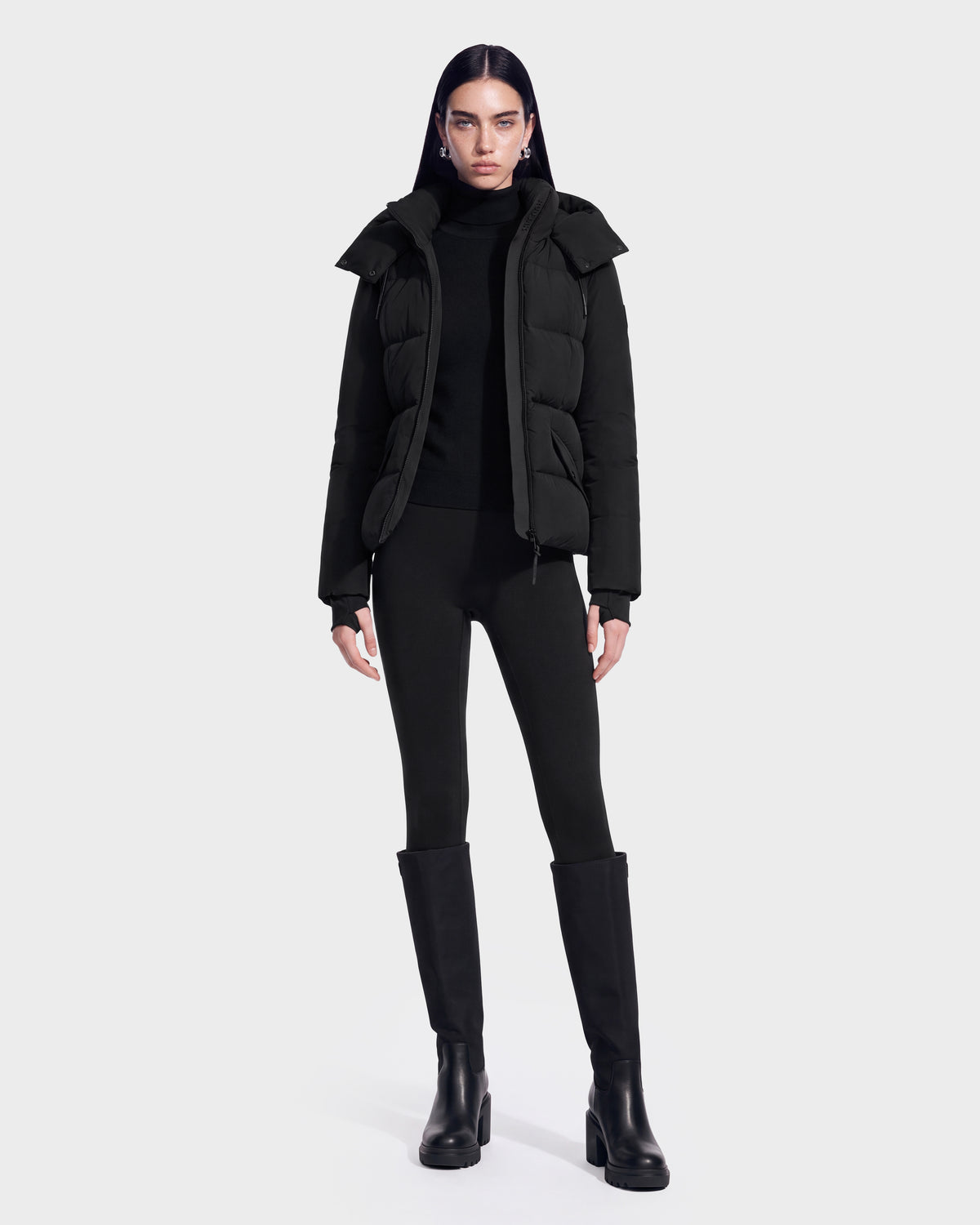 A person stands facing forward, wearing the Rudsak ADELINE - 8124544 Black women’s down puffer jacket made with RDS-certified down, paired with a black turtleneck, black leggings, and tall black boots against a plain white background.