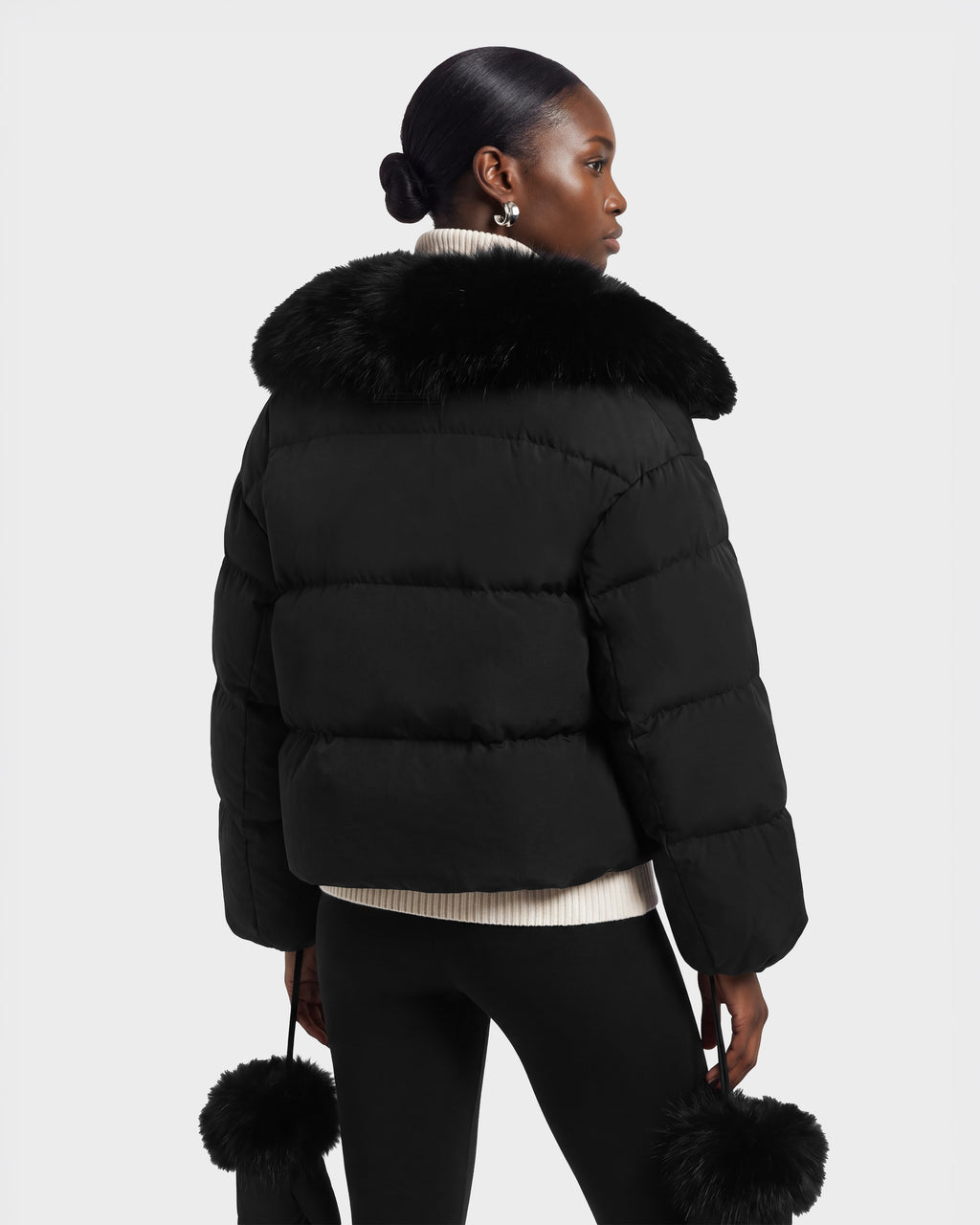 A woman wears a black Rudsak ALONDRA - 8126992 cropped puffer jacket with a large fur collar and matching fur-trimmed cuffs.