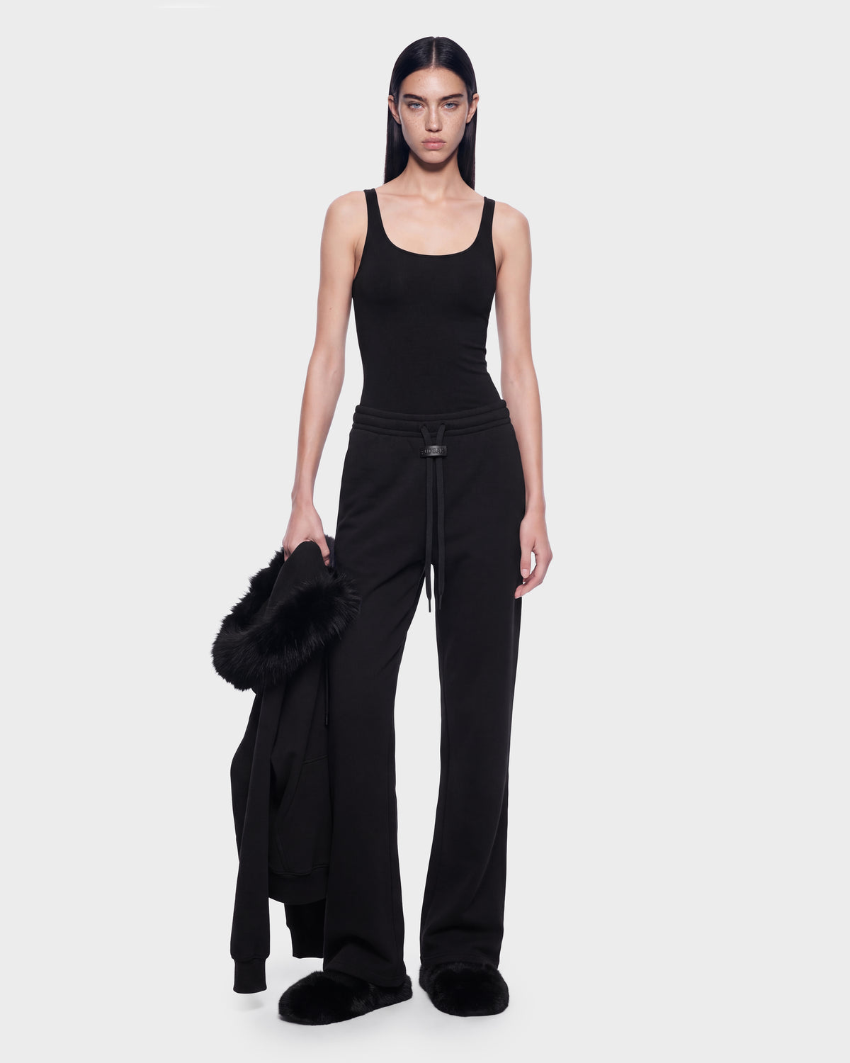 A person wearing a black tank top and refined off-duty black sweatpants with black slippers holds a black ANGEL - 8526725 Black jacket with a fur-trimmed hood.