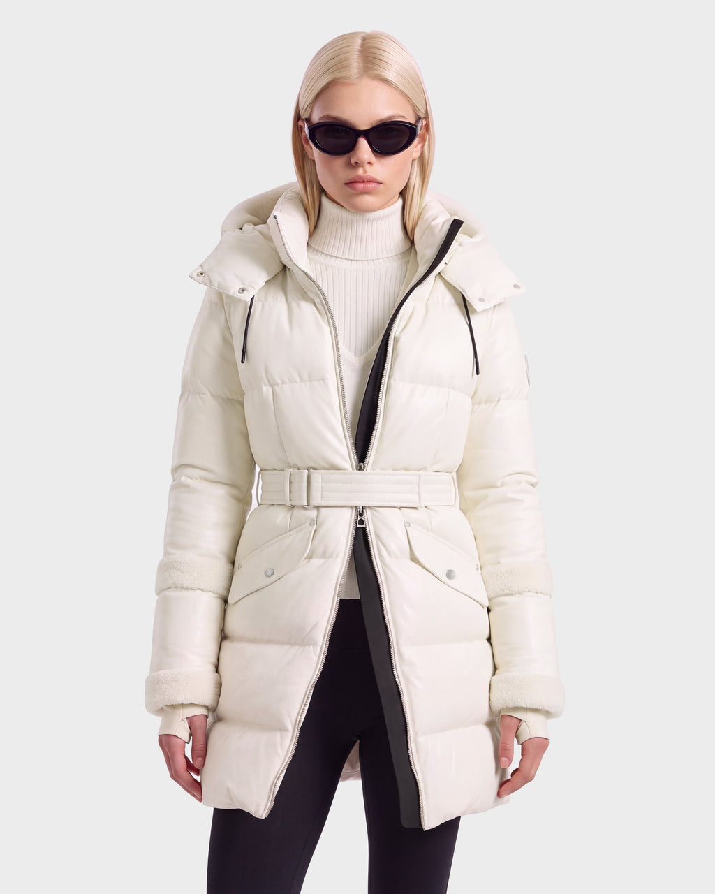 A person wearing a knee-length, cream-colored Rudsak AUDREY L - 8124585 Vanilla down puffer jacket with a buckled belt over a white turtleneck sweater and black leggings.