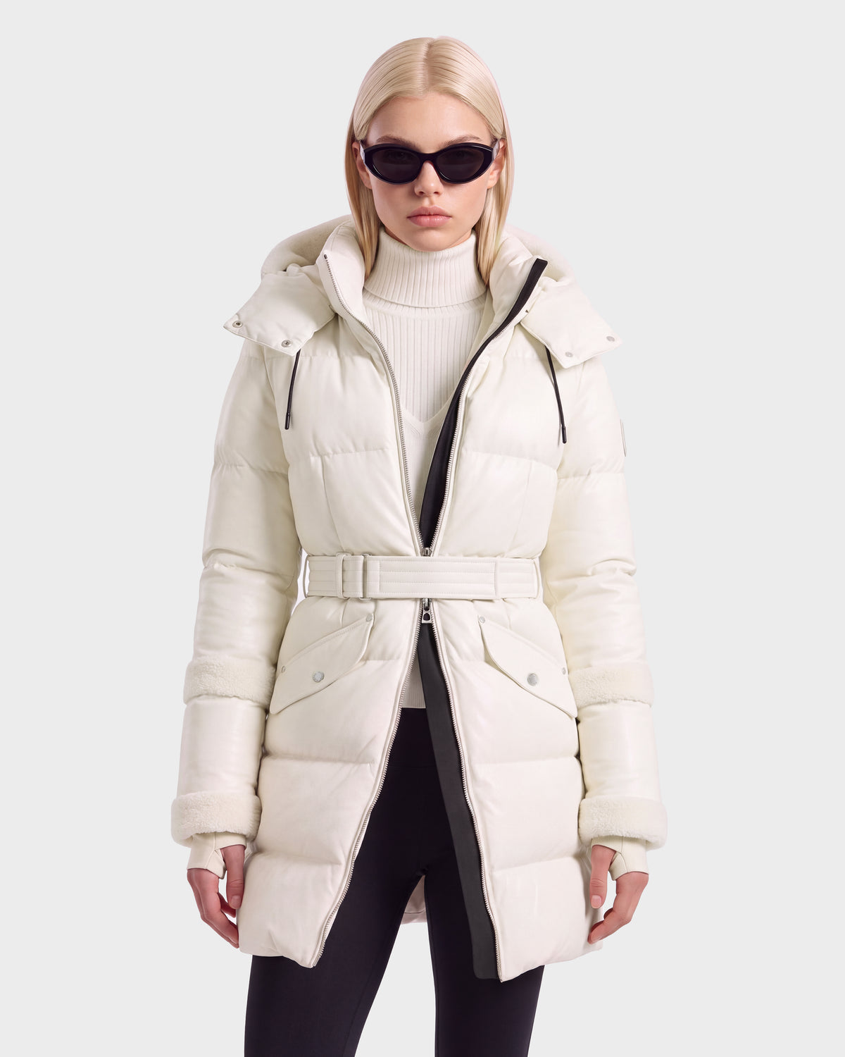 A person wearing a knee-length, cream-colored Rudsak AUDREY L - 8124585 Vanilla down puffer jacket with a buckled belt over a white turtleneck sweater and black leggings.