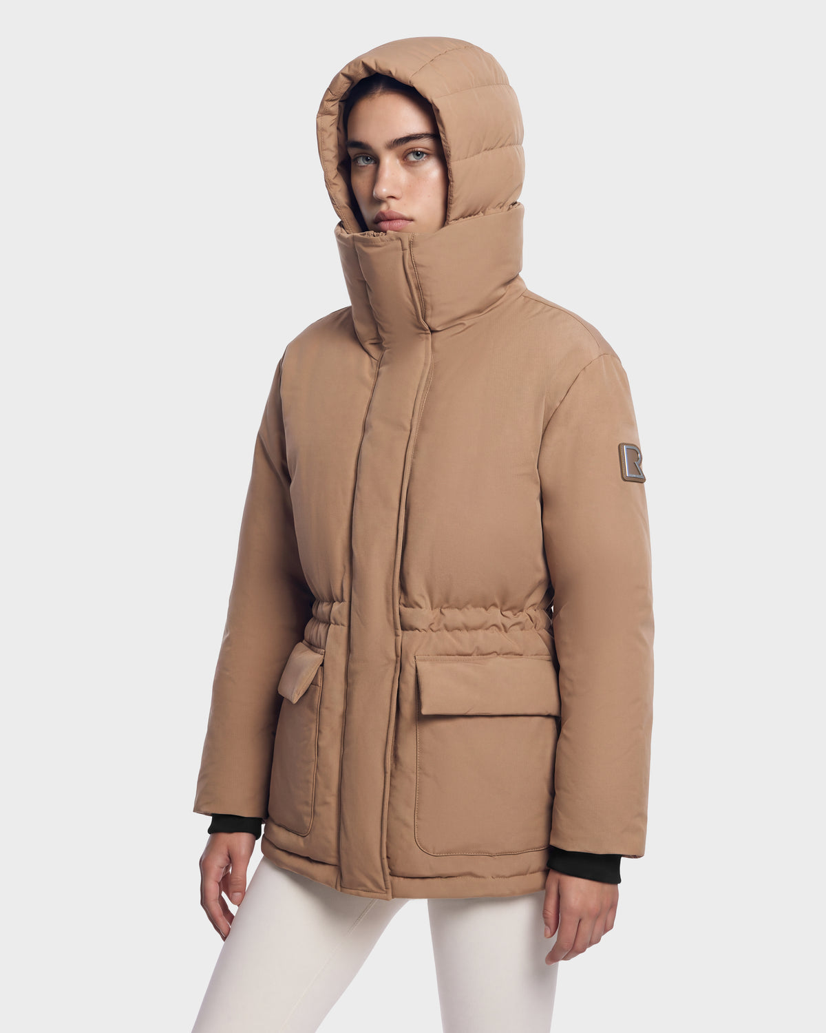 A model wears a camel-colored BHIMA - 8126515 Rudsak hooded puffer jacket with a cinched waist and two large front flap pockets.