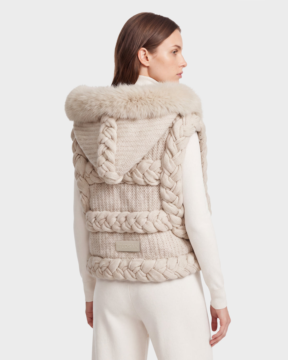 A woman with straight brown hair, seen from behind, wears the CICELY - 8526686 Vanilla knit vest by Rudsak—featuring a cream color with bold braided details and a cozy faux fur-trimmed hood—layered over matching long-sleeve top and pants for a perfect winter look.