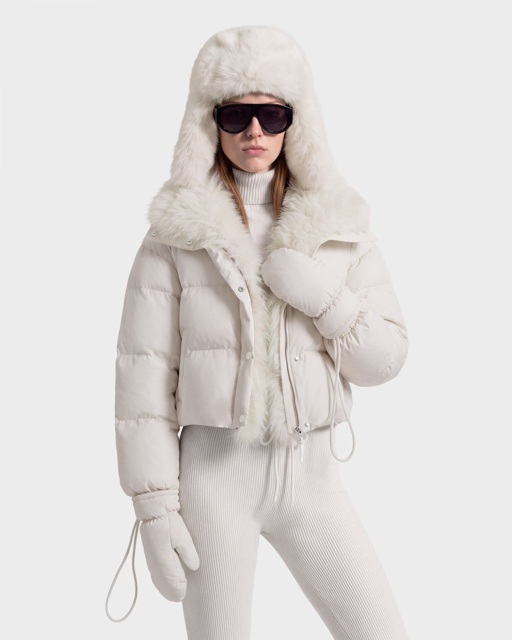 A person wearing an ALONDRA - 8126992 Vanilla white, cropped puffer jacket with a faux fur-lined hood, matching mittens, and sunglasses.