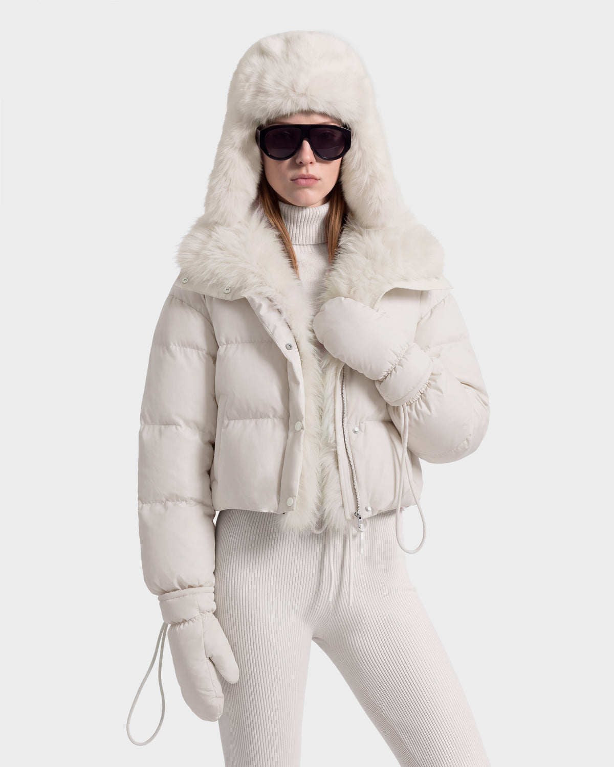 A person wearing an ALONDRA - 8126992 Vanilla white, cropped puffer jacket with a faux fur-lined hood, matching mittens, and sunglasses.