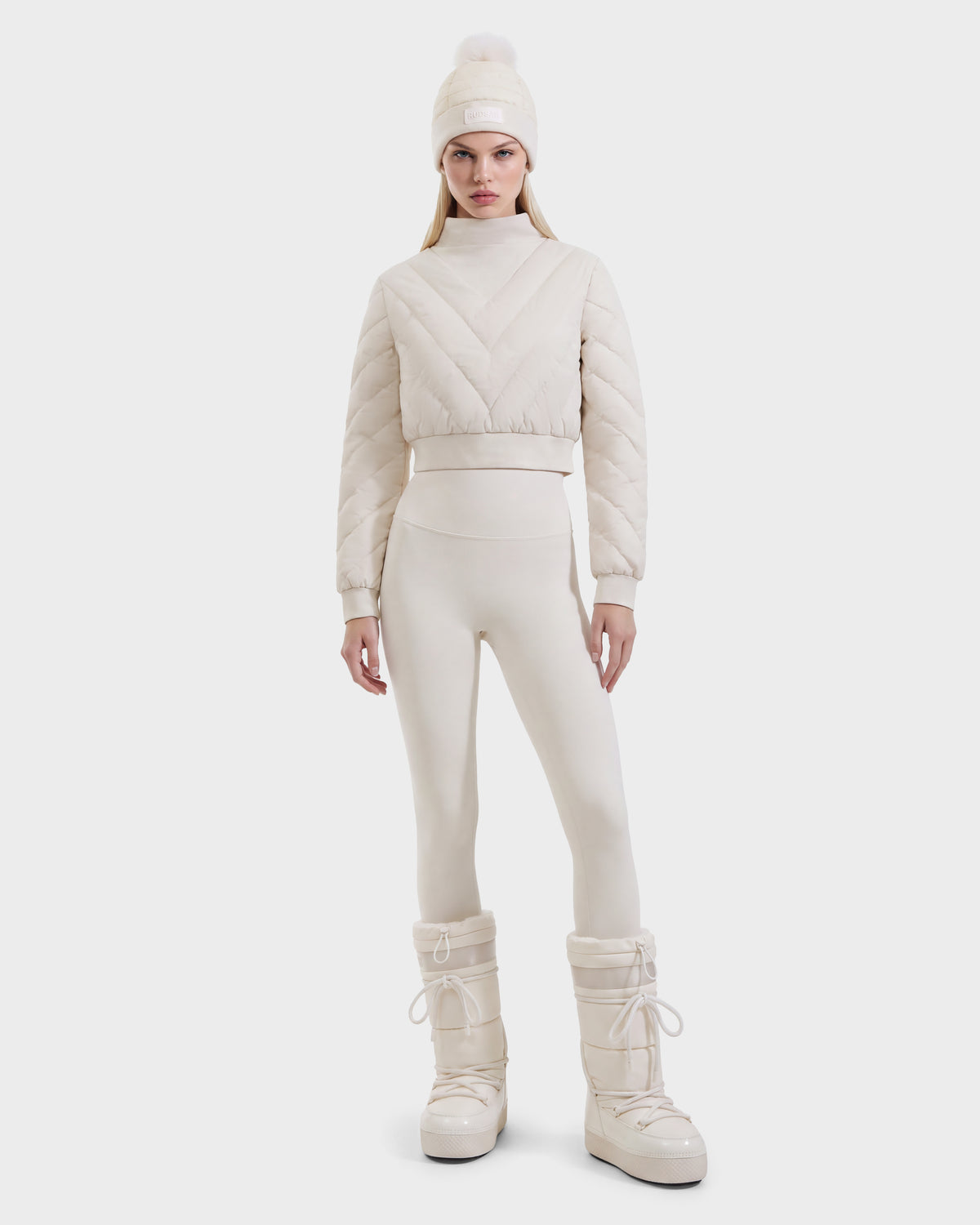 A model wearing a KATYA - 8526531 Vanilla quilted RDS-certified down crop top, leggings, matching beanie with a pompom, and tall lace-up winter boots.