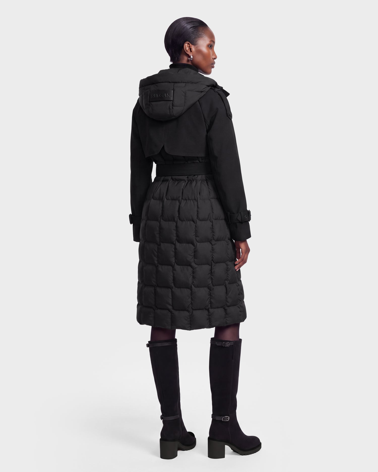 A person with short hair is shown from the back, wearing the Rudsak ELVIE - 8126512 Black knee-length quilted coat with a hood and high black boots against a light gray background.