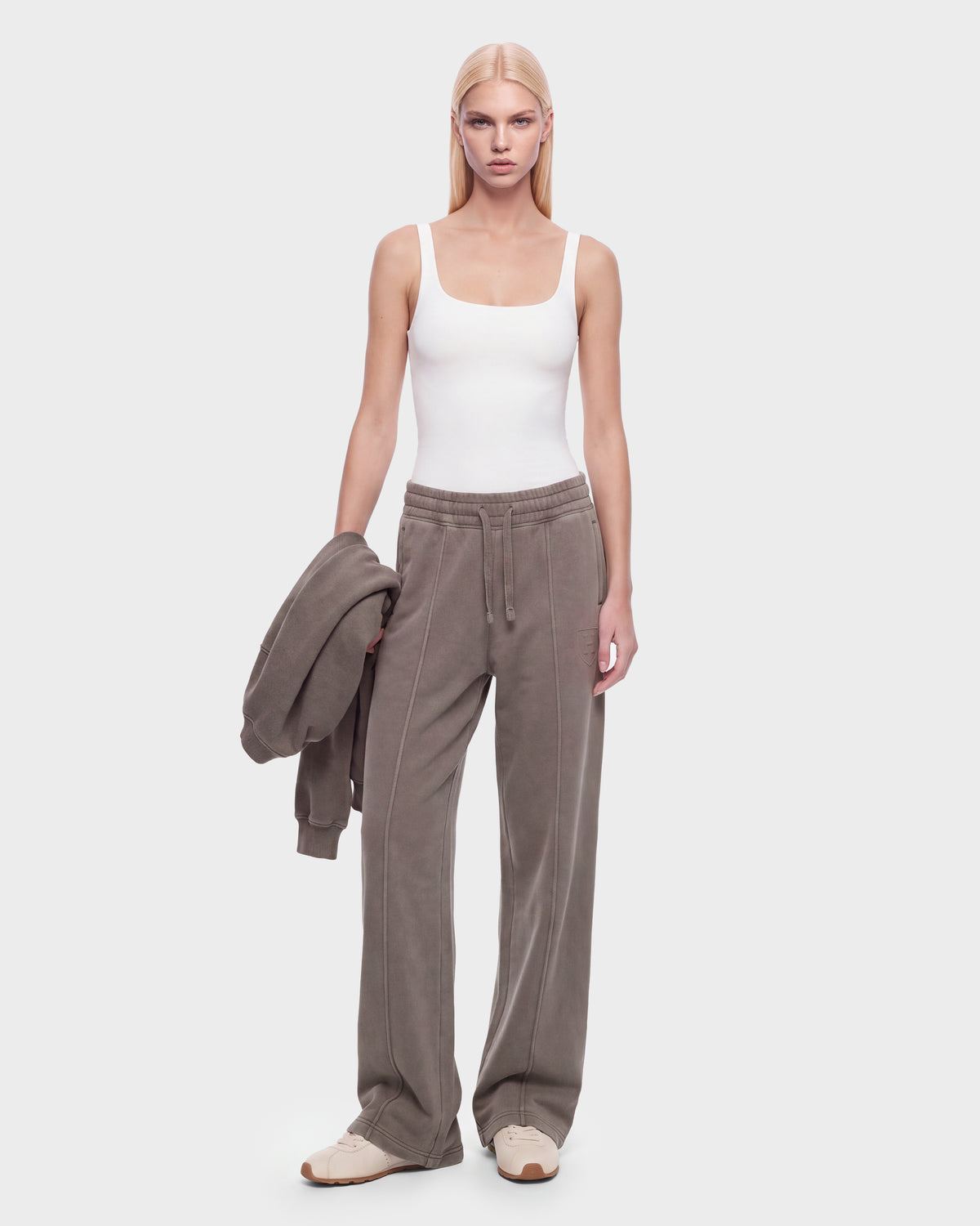 A woman with straight blonde hair wears a white tank top, gray heavyweight jersey jogger pants, and Rudsak’s GAELLE HV - H526902 Taupe sneakers, holding a matching gray jacket in one hand while standing against a plain white background.
