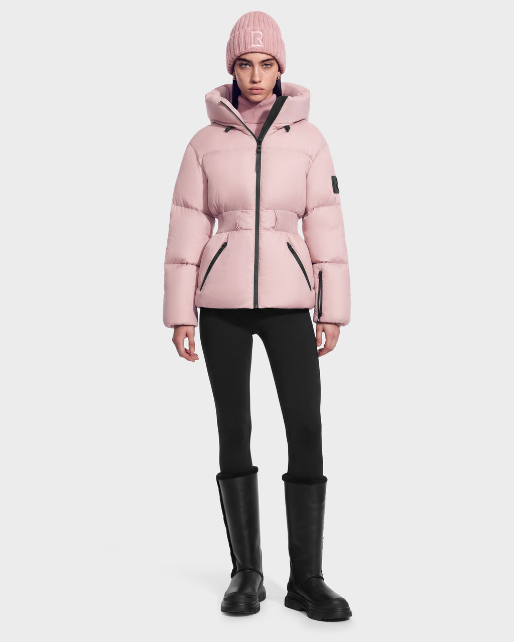 A person wears a light pink Rudsak DIAMOND - 8126772 Vintage Rose quilted puffer jacket with a belted waist, a matching knit beanie, black leggings, and black knee-high boots for winter warmth.