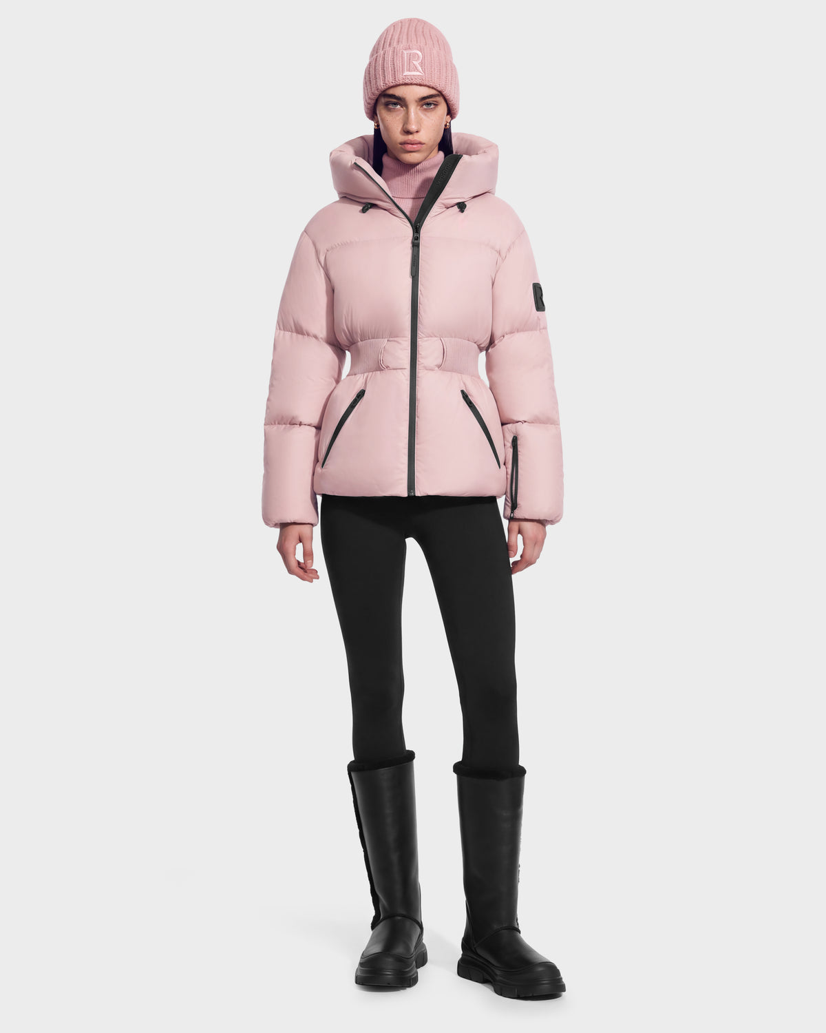 A person wears a light pink Rudsak DIAMOND - 8126772 Vintage Rose quilted puffer jacket with a belted waist, a matching knit beanie, black leggings, and black knee-high boots for winter warmth.