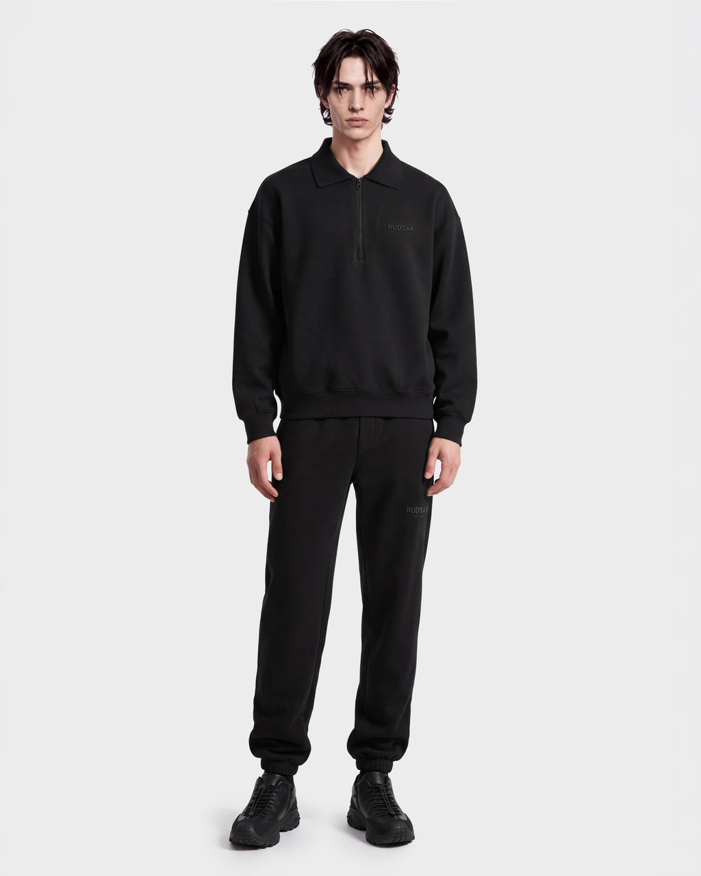 A person faces forward wearing the Rudsak IDRIS - 6526500 Black polo collar sweater with matching sweatpants and sneakers, presenting a relaxed look against a plain white background.