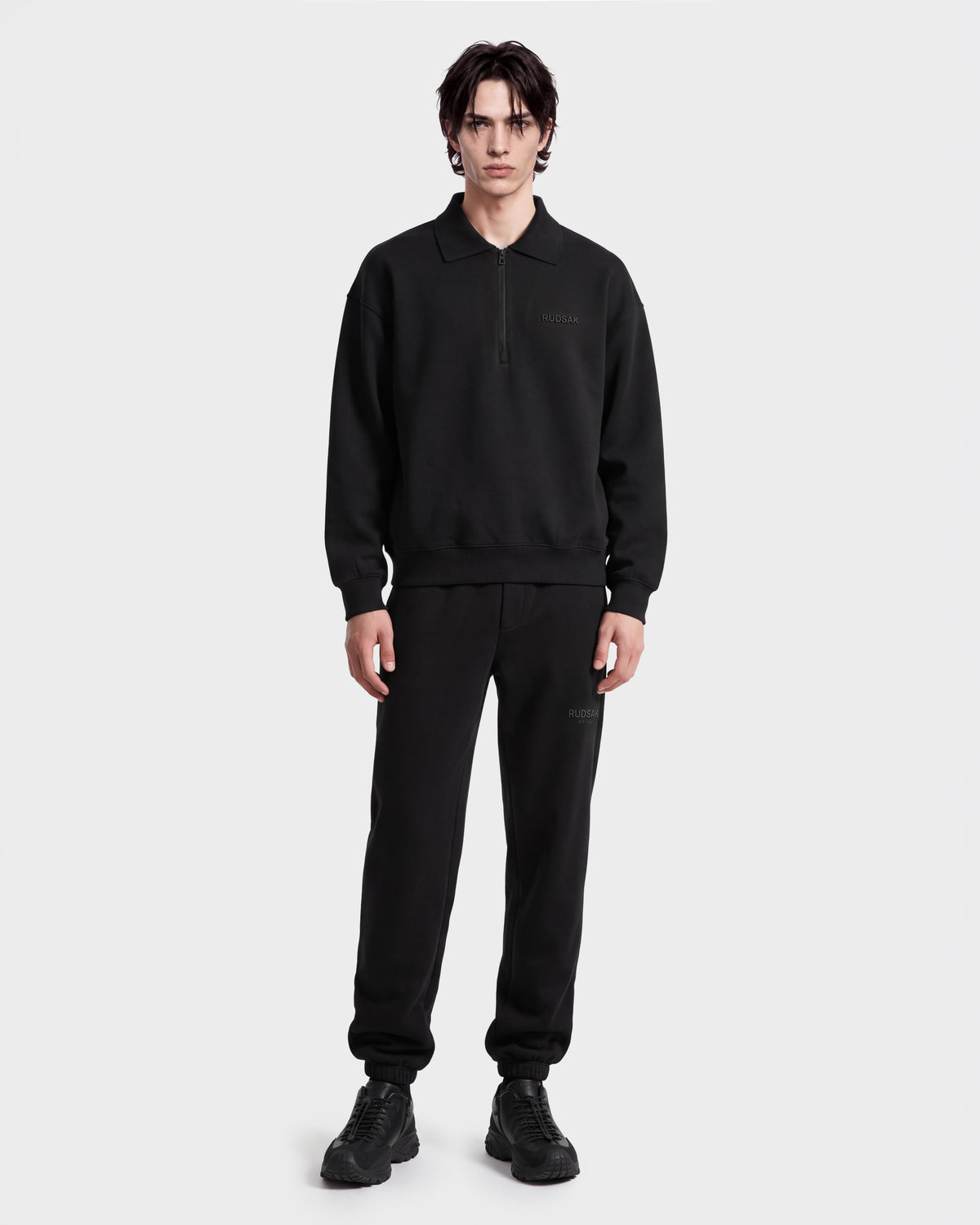 A person faces forward wearing the Rudsak IDRIS - 6526500 Black polo collar sweater with matching sweatpants and sneakers, presenting a relaxed look against a plain white background.