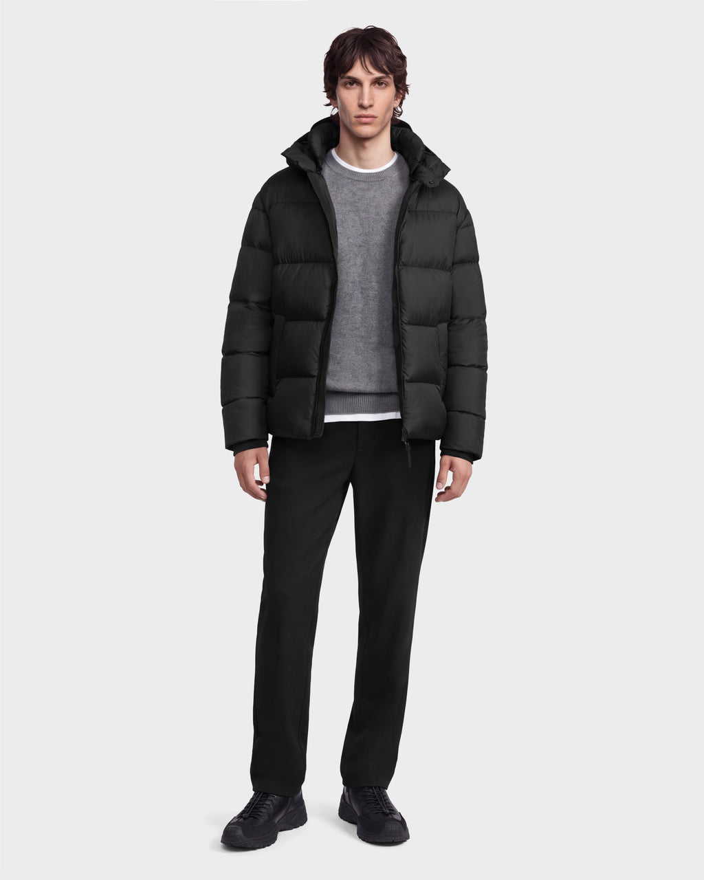A man wearing a black Rudsak JASON - 6126803 Black puffer jacket with a hood over a grey sweater, black trousers, and black sneakers.