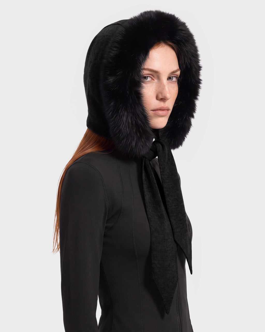 A person with long hair wears a black outfit and the KARYMA - 8426507 Black hood-and-scarf hybrid by Rudsak, featuring plush trim, while looking slightly to the side against a plain light background—an ideal choice for those seeking chic winter accessories.