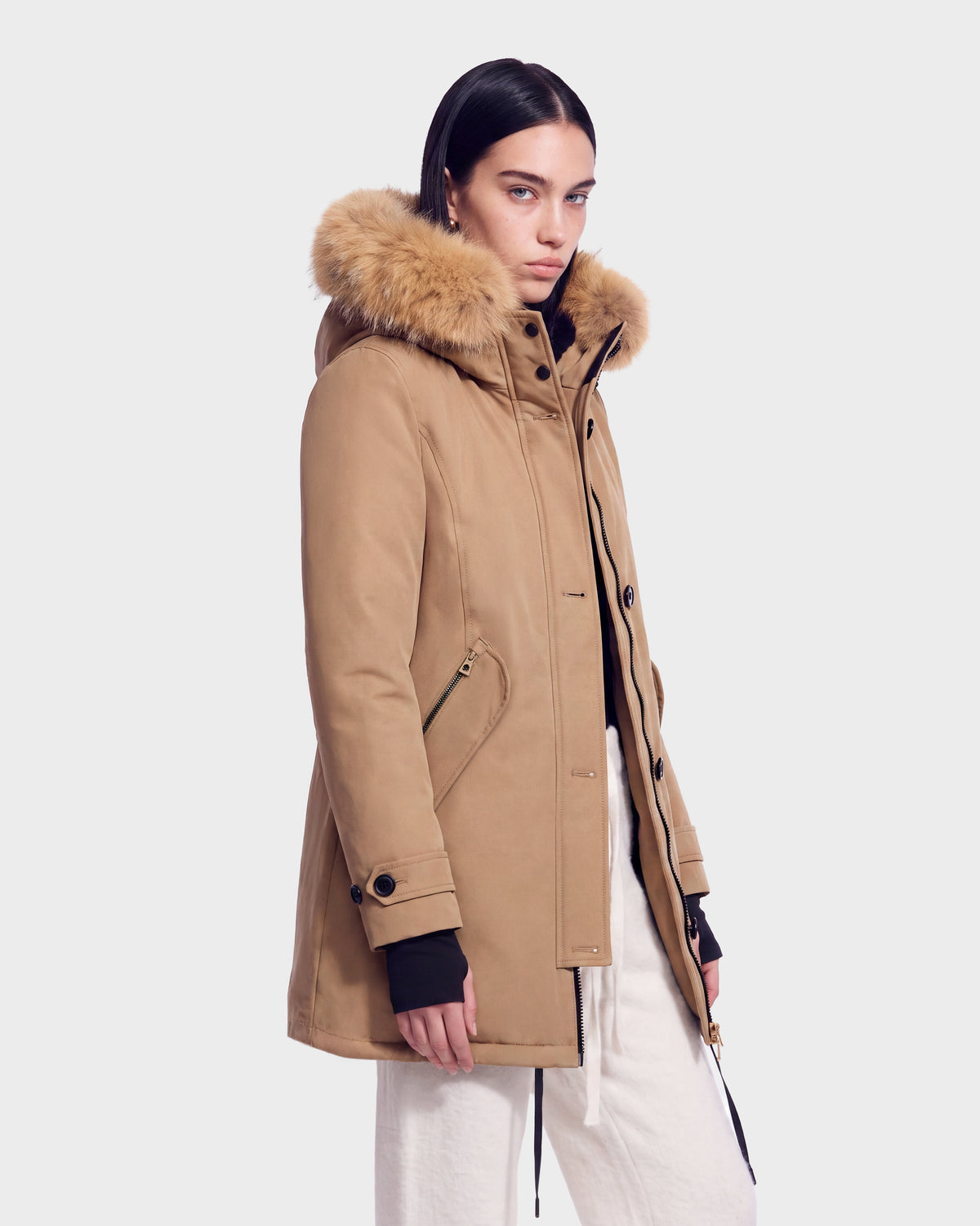 A person stands against a plain light background, wearing the Rudsak KEYSHIA - 8126517 Camel insulated jacket with fur-lined hood, black shirt sleeves at the cuffs, and light-colored pants.