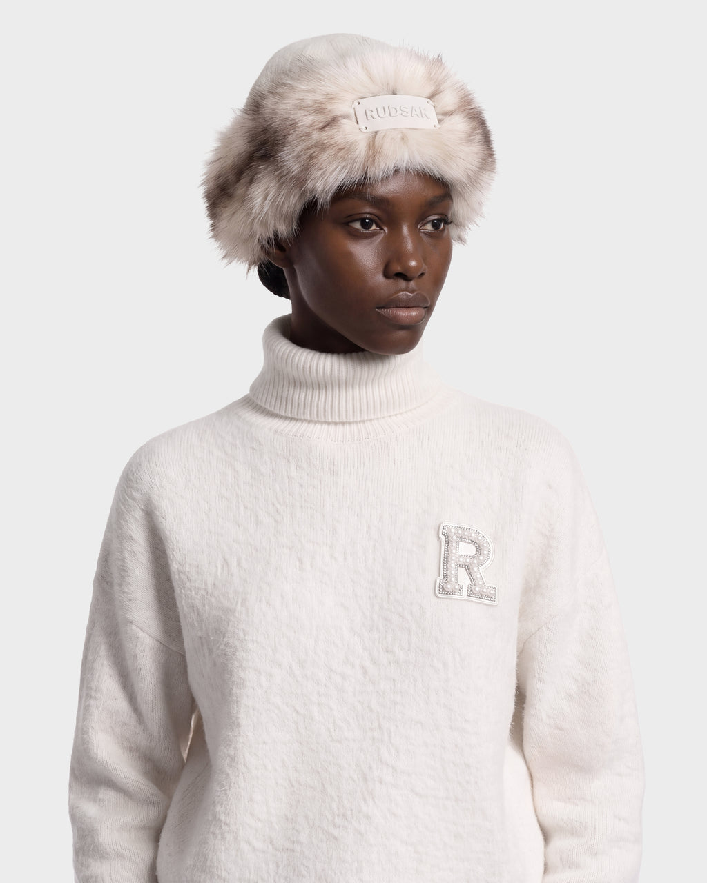 A model wears a cream-colored LORETTA W - 8426510 Vanilla turtleneck sweater with a decorative letter R patch and a matching RUDSAK faux fur-trimmed bucket hat.