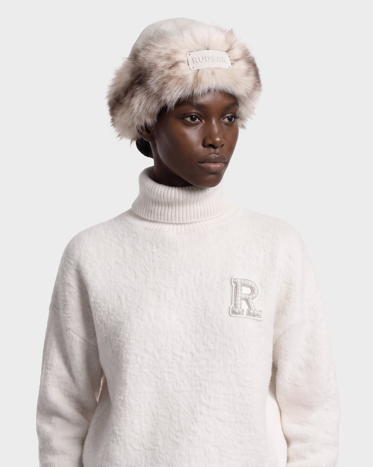 A model wears a cream-colored LORETTA W - 8426510 Vanilla turtleneck sweater with a decorative letter R patch and a matching RUDSAK faux fur-trimmed bucket hat.