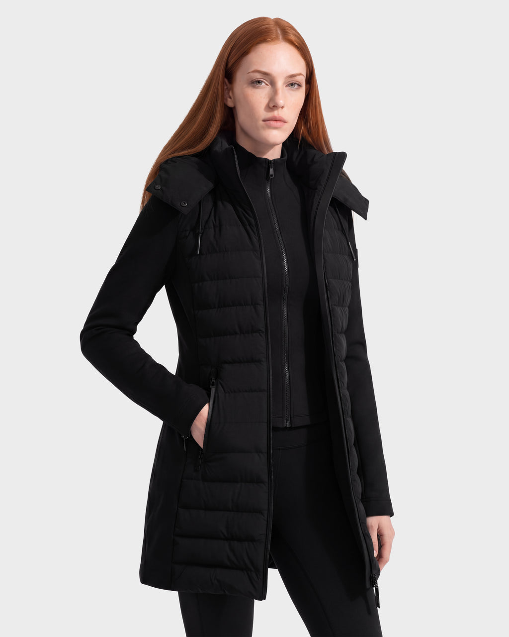 A woman with long red hair wears the Rudsak MERINA - 8125056 Black quilted down jacket, featuring a zipper and side pockets, as she stands against a plain light gray background.