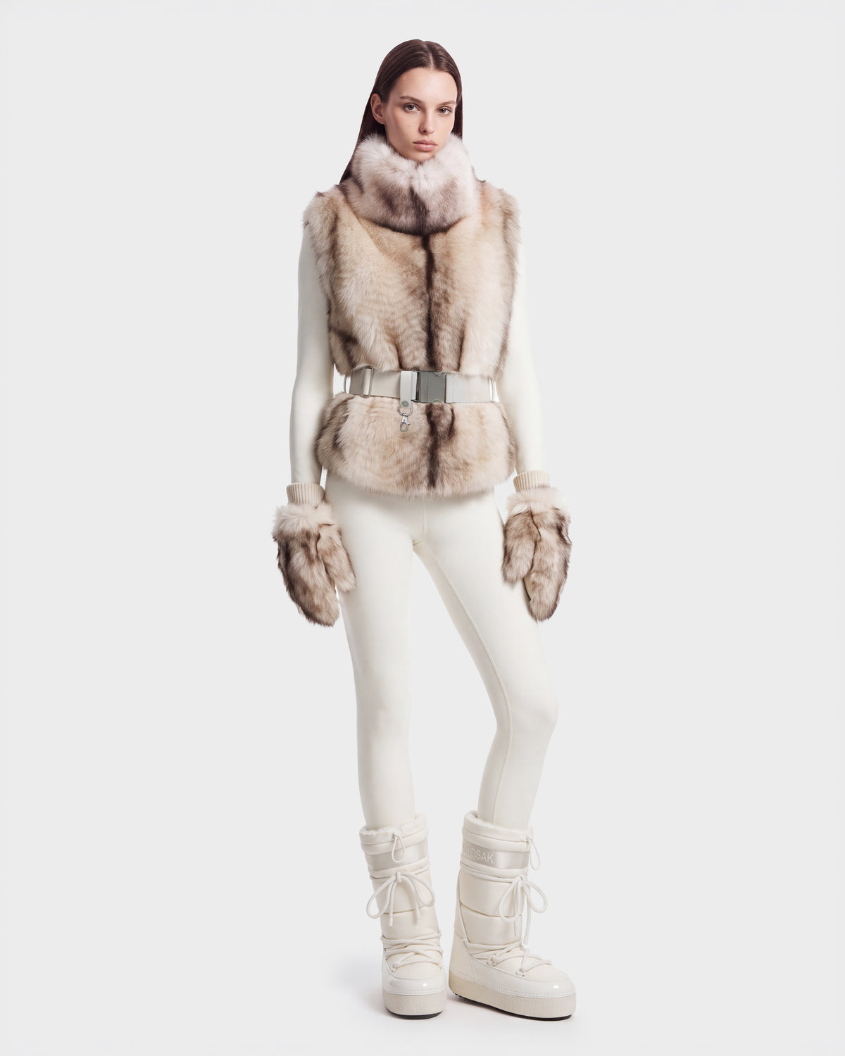 A woman wears a MAULIN - 8524611 Mix Vanilla faux fur vest with a silver-buckled belt over a white mock-neck pullover and leggings, finished with matching fur mittens and white insulated boots.