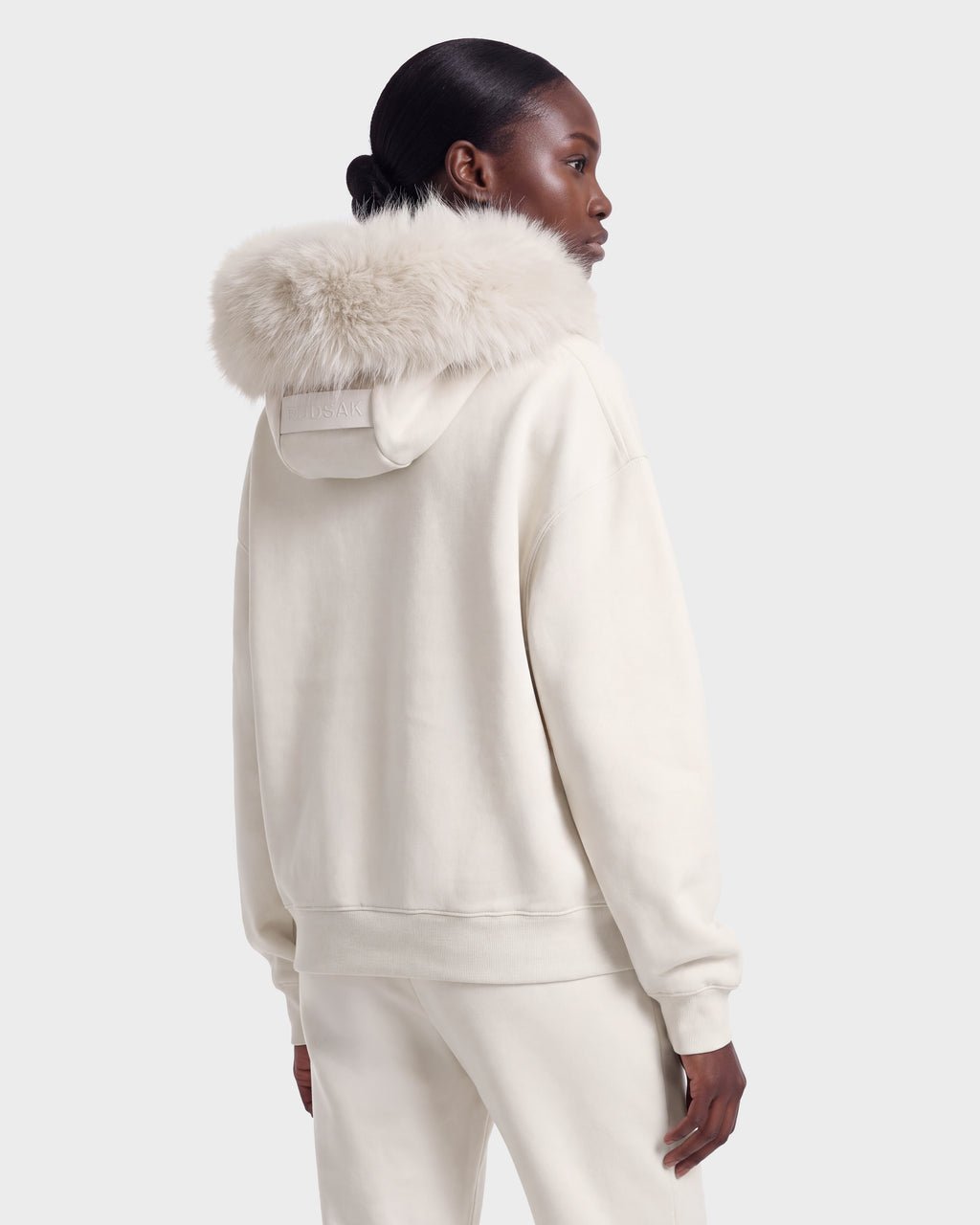 Someone wears the MILENA - 8526689 Vanilla hoodie by Rudsak, featuring a large, fluffy fur-lined hood, standing with their back to a plain background.