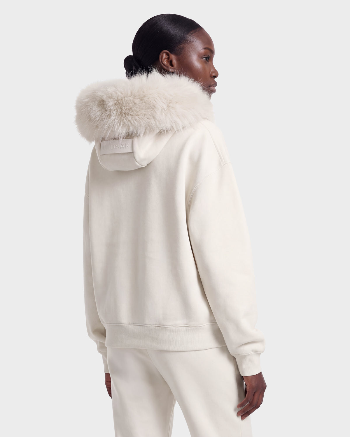 Someone wears the MILENA - 8526689 Vanilla hoodie by Rudsak, featuring a large, fluffy fur-lined hood, standing with their back to a plain background.