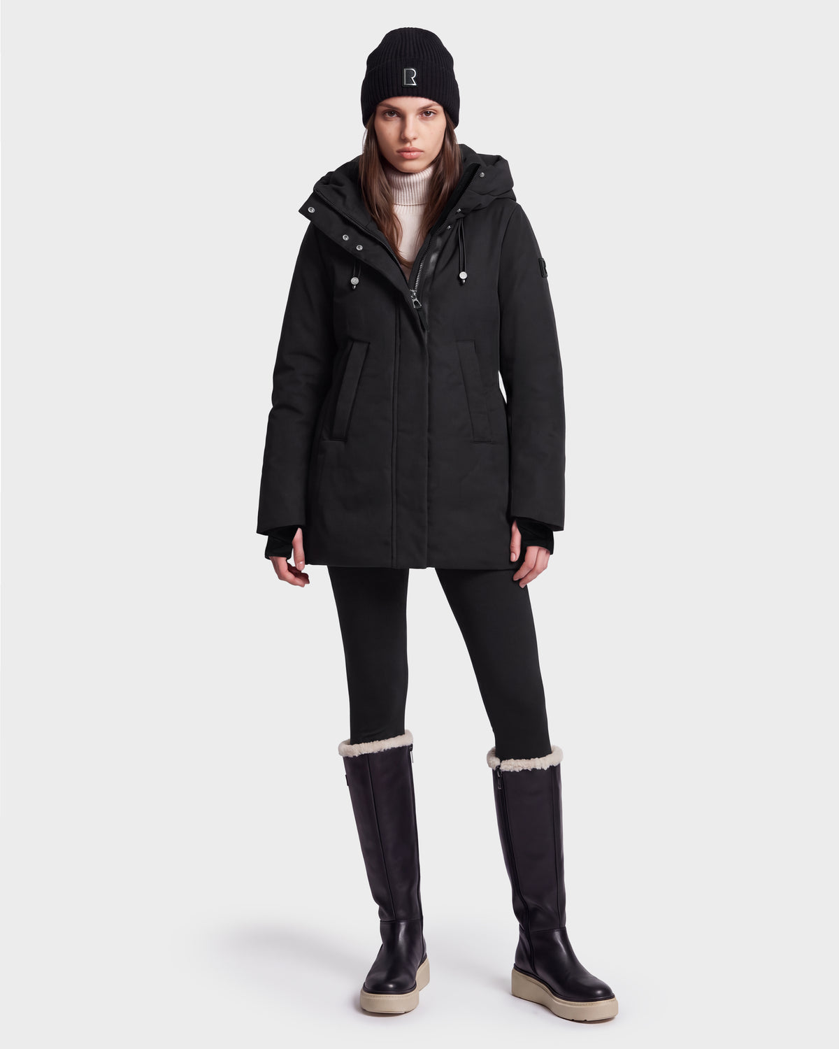 A person wearing the Rudsak OPAL - 8126618 Black knee-high boots with white soles, along with a black beanie, black parka, and black leggings, stands against a plain white background.