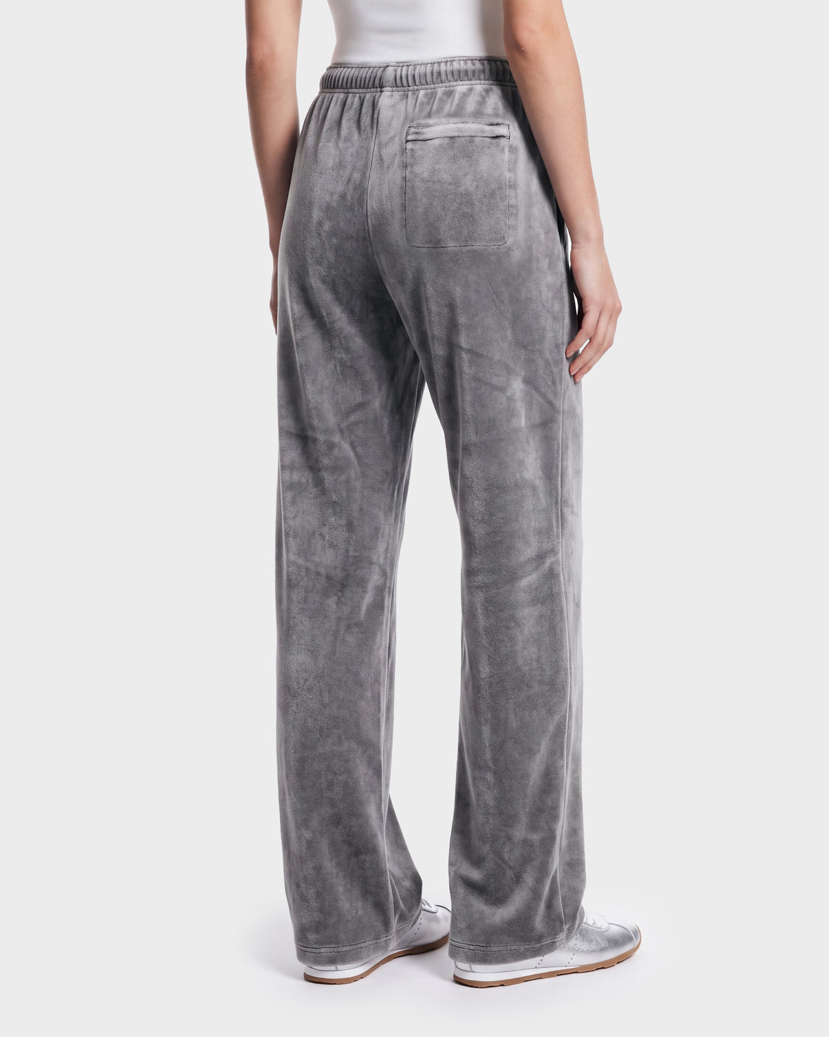 A person is shown from the back against a plain background, wearing the Rudsak OLIVYA HV - H526903 Titanium pants—loose-fitting gray velour pants featuring an elastic waistband and a back pocket—paired with white sneakers and a white top.