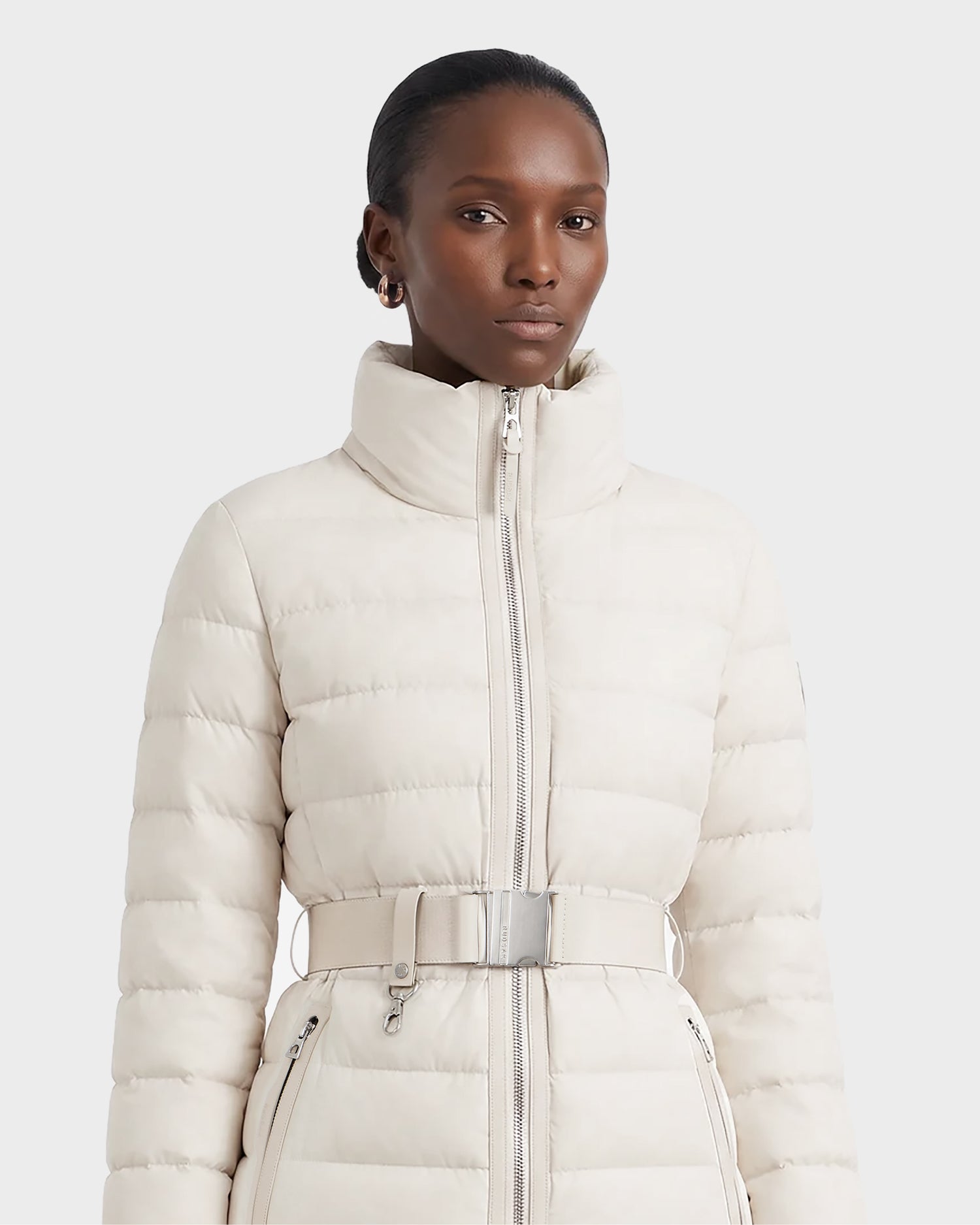 A woman wearing the Rudsak PELIN S - 8124813 quilted jacket in Vanilla, featuring a belted waist and high collar, stands against a plain light background, looking forward with a neutral expression.