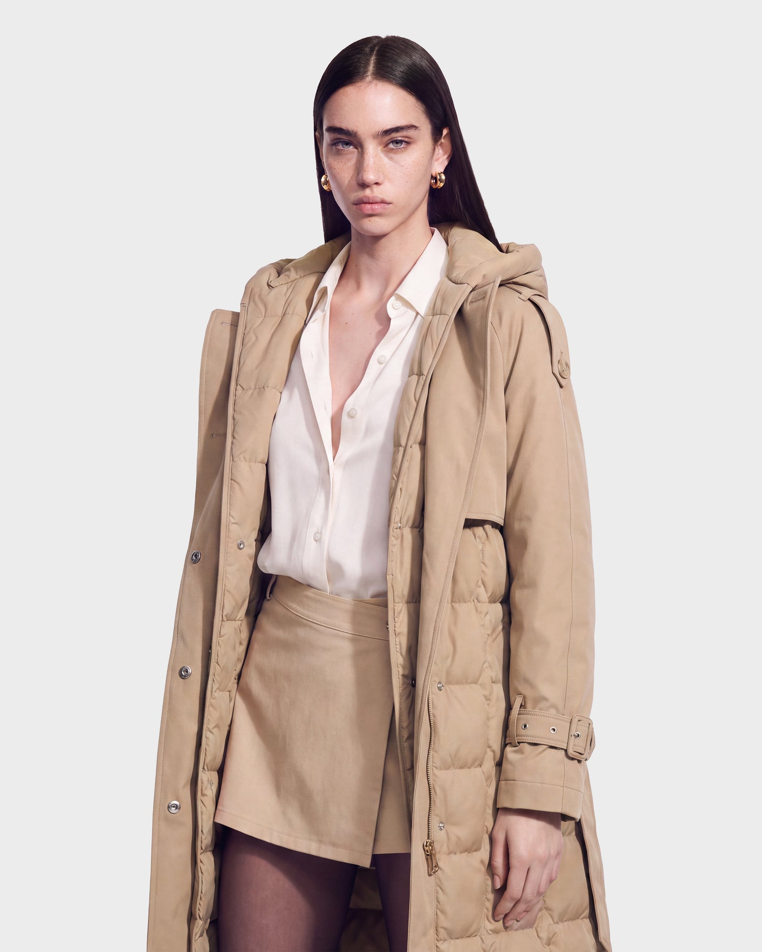 A woman with straight brown hair wears the Rudsak ELVIE - 8126512 Almond trench coat, which is RDS-certified down in a beige hue, layered over a white shirt and beige skirt. She stands against a plain background, looking forward with a neutral expression.