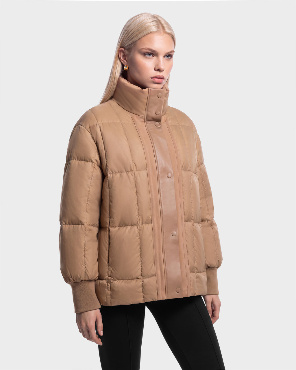 A person wearing a camel-colored Rudsak ROSALINDA - 8126523 Camel boxy puffer jacket with a high collar, snap-button closure, and ribbed cuffs.