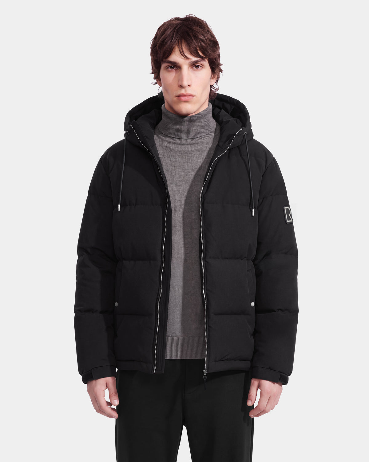 A person with short brown hair is wearing the Rudsak STERLING EL - 6126759 Black hooded puffer jacket over a gray turtleneck sweater and black pants, standing against a plain white background.