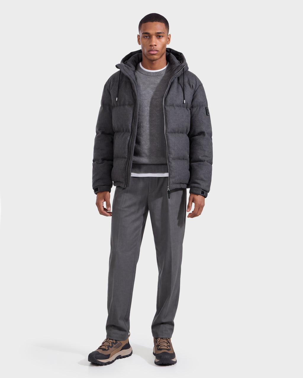 A man wearing the Rudsak STERLING W - 6126760 Heather Charcoal puffer jacket over a gray sweater and shirt, paired with gray pants and brown sneakers, stands facing forward against a plain white background.