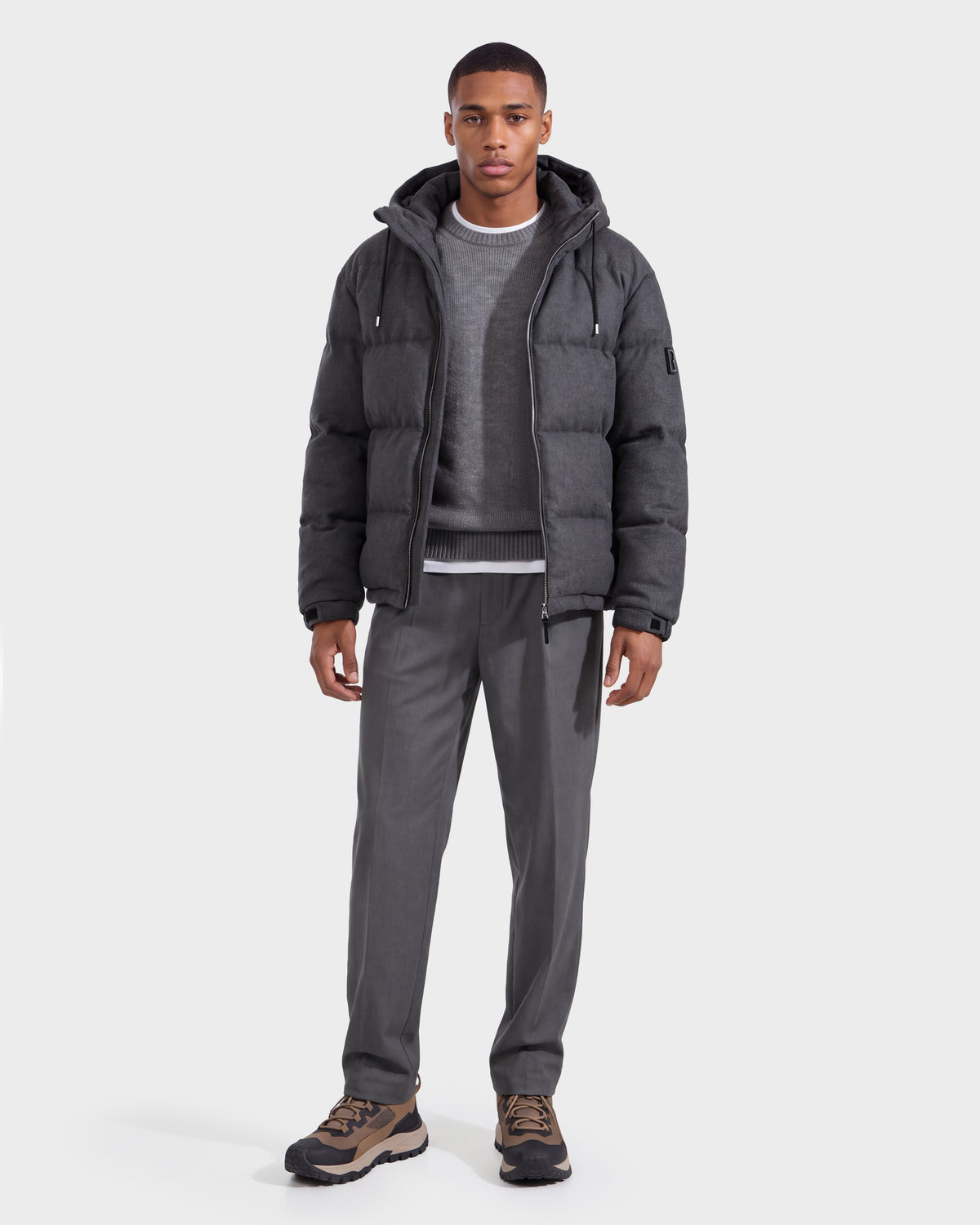 A man wearing the Rudsak STERLING W - 6126760 Heather Charcoal puffer jacket over a gray sweater and shirt, paired with gray pants and brown sneakers, stands facing forward against a plain white background.