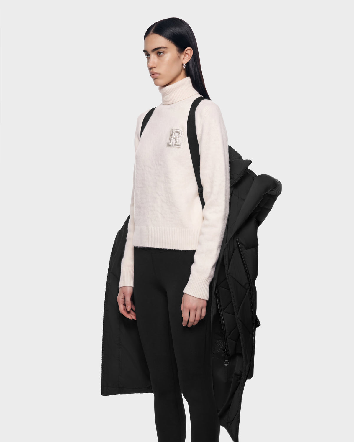 A person wearing a cream-colored turtleneck sweater with an R patch and black leggings, with a TAYLOR M - 8125965 Black puffer coat by Rudsak draped over their shoulders.