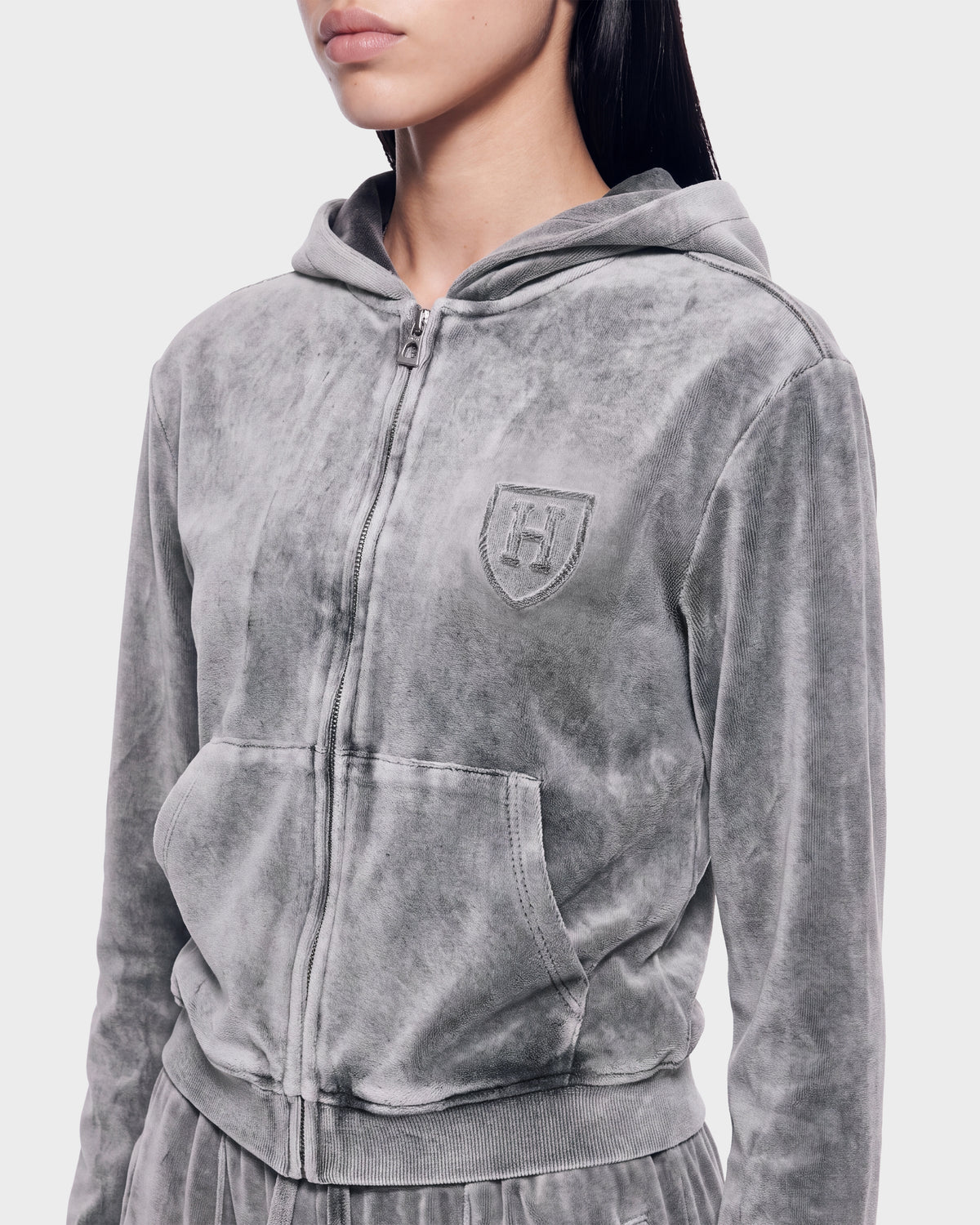A person wearing the Rudsak TYRA HV - H526904 Titanium velour zip-up hoodie in light gray, featuring a front pocket and embroidered crest, is shown from the shoulders to the waist against a plain background.