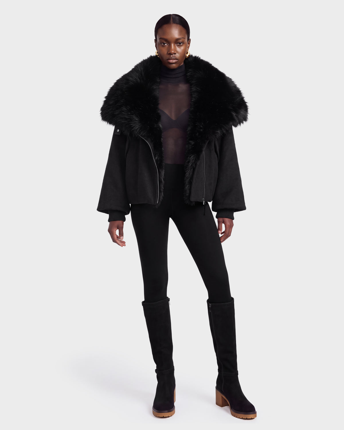 A person wearing the Rudsak ZARIA - 8126999 Black fur-collared jacket stands against a plain white background, styled with a sheer black top, black leggings, and knee-high black boots.