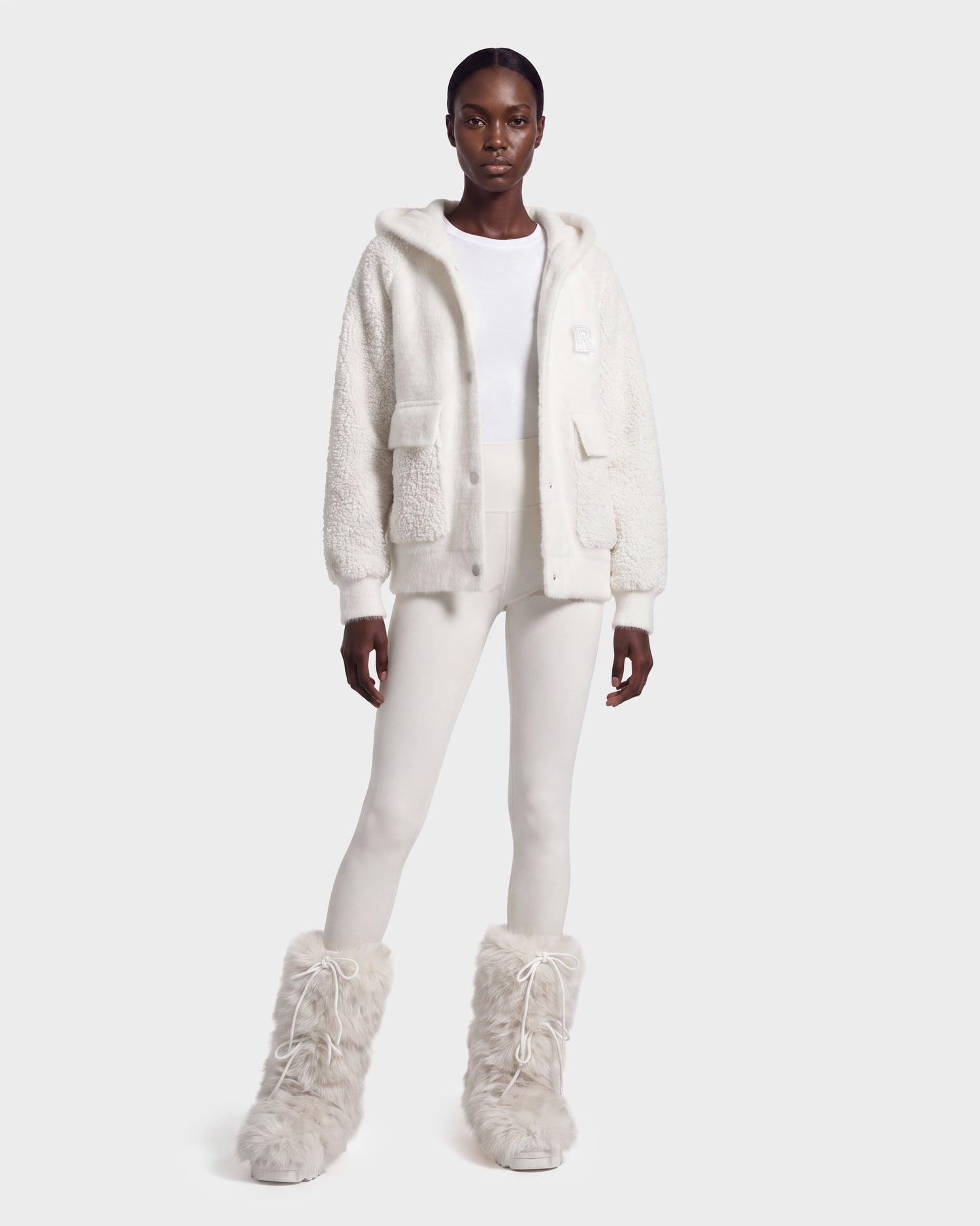 A woman stands against a plain light background wearing the Rudsak DAKOTA - 8526515 Iceland white fuzzy hooded jacket, paired with a white top, white pants, and tall white furry boots.