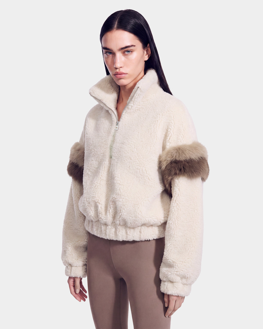 A woman is seen wearing the Rudsak ALEIA - 8526693 Vanilla faux-shearling zip-up jacket with fur accents on the sleeves, paired with taupe leggings, as she stands against a plain, light background.