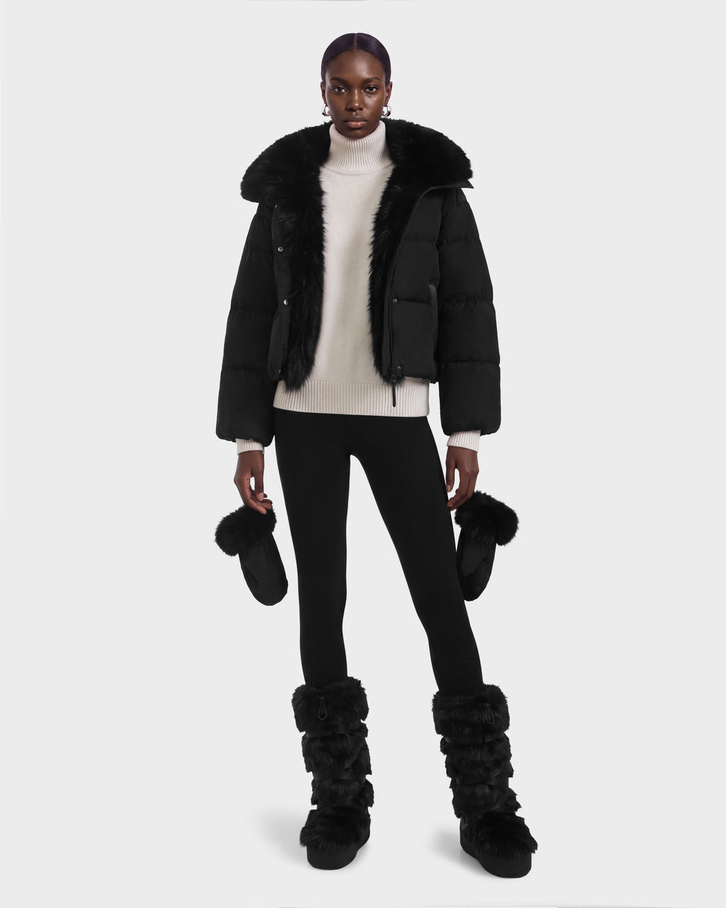 A model wears a black Rudsak ALONDRA - 8126992 Black puffer jacket with a fur-trimmed collar, a white turtleneck sweater, black leggings, and matching black faux fur mittens and tall boots.