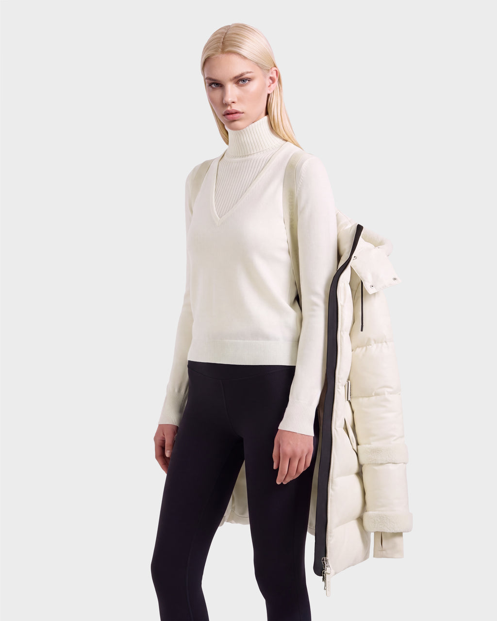 A person wearing a white V-neck sweater over a white turtleneck and black leggings, with a Rudsak AUDREY L - 8124585 Vanilla knee-length down puffer jacket draped over their shoulders.