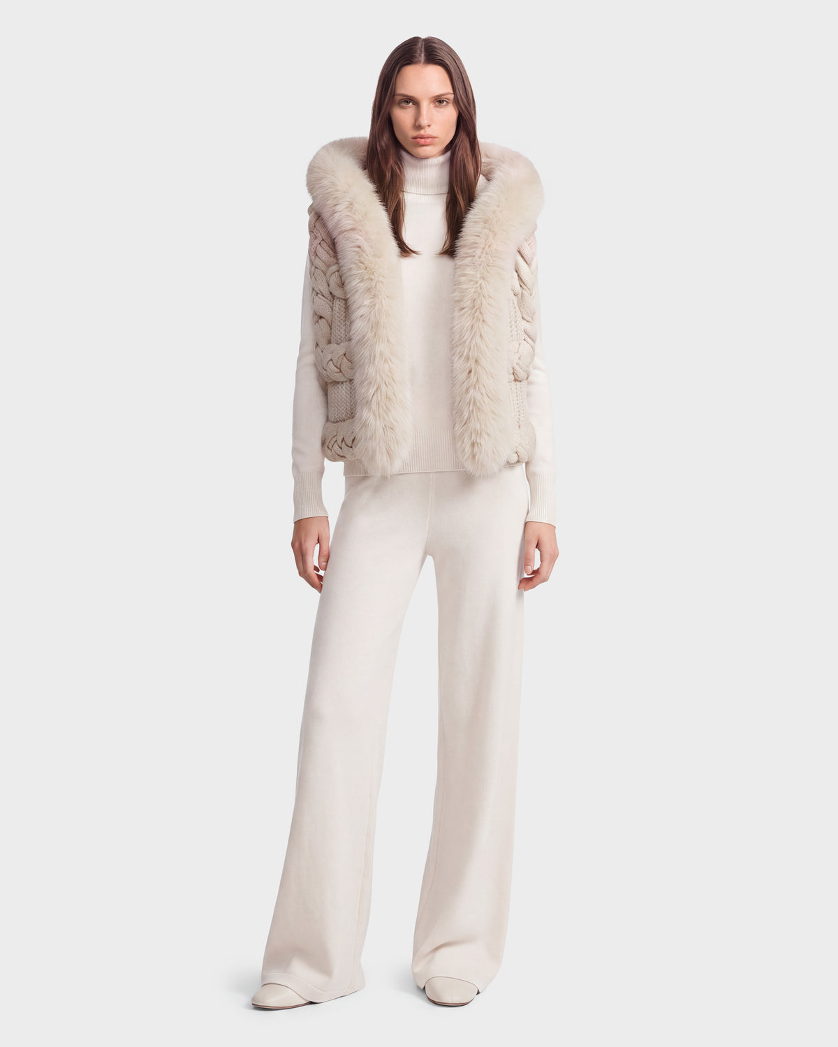 A woman stands against a plain light background wearing the Rudsak CICELY - 8526686 Vanilla—a cream turtleneck sweater featuring a sleeveless knit vest with a faux fur-trimmed hood—paired with wide-legged pants, creating an ensemble perfect for winter layering.