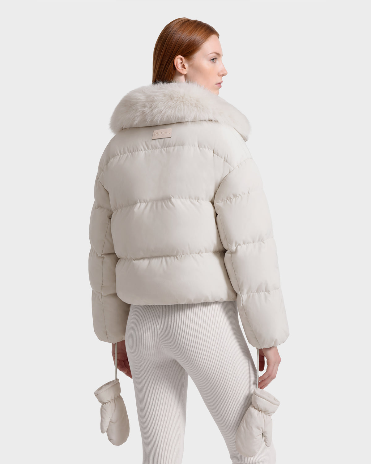 Woman seen from the back wearing a cream-colored Rudsak ALONDRA - 8126992 Vanilla cropped puffer jacket with a fur-trimmed collar and matching mittens.