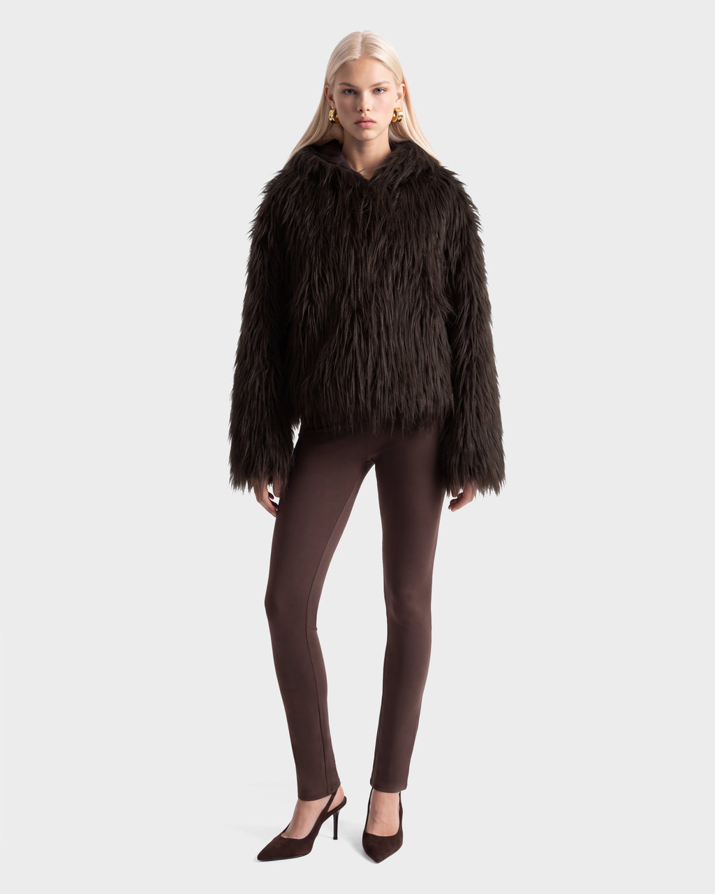 A model wearing a dark brown, plush faux fur ELLISON - 8525678 Espresso hoodie jacket, brown leggings, and pointed-toe heels.