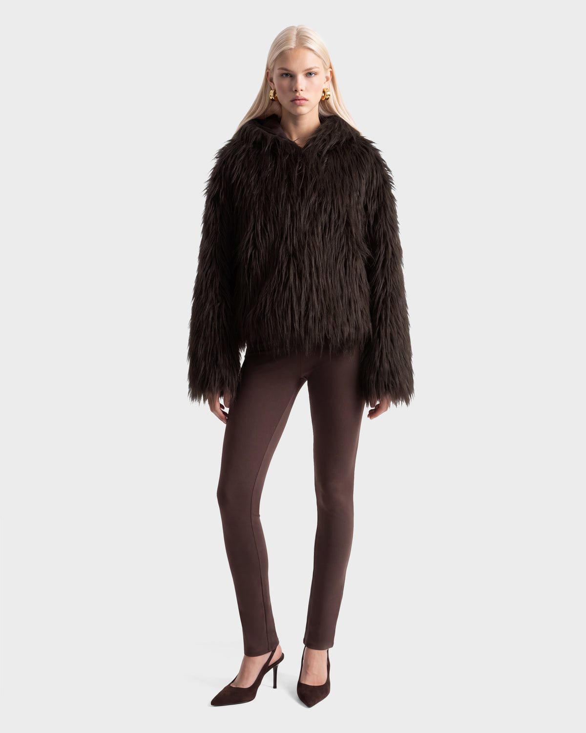 A model wearing a dark brown, plush faux fur ELLISON - 8525678 Espresso hoodie jacket, brown leggings, and pointed-toe heels.