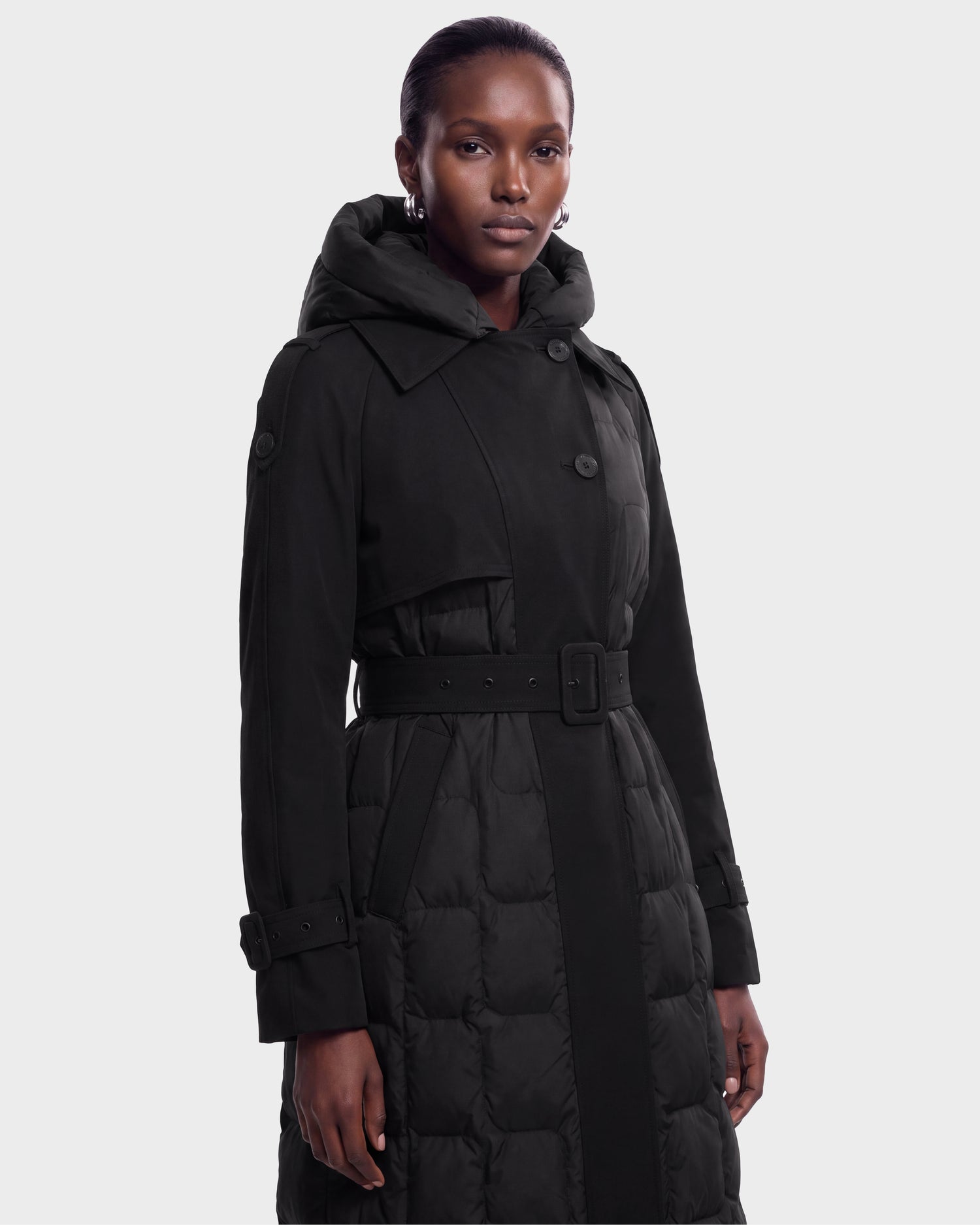 A person is wearing the Rudsak ELVIE - 8126512 Black belted trench coat with a hood, standing against a plain light gray background and looking directly at the camera with a neutral expression.