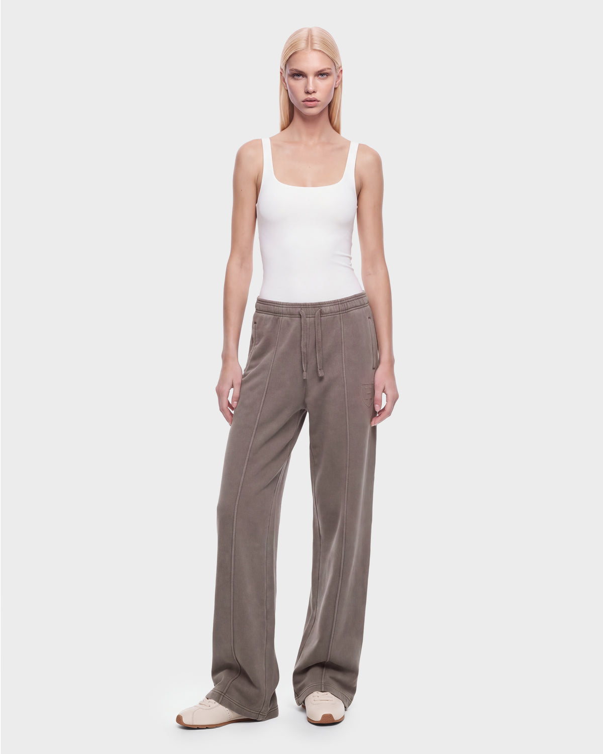 A person with straight blonde hair wears a fitted white tank top, loose brown heavyweight jersey jogger pants with front seams, and Rudsak’s GAELLE HV - H526902 Taupe sneakers, standing against a plain light gray background.