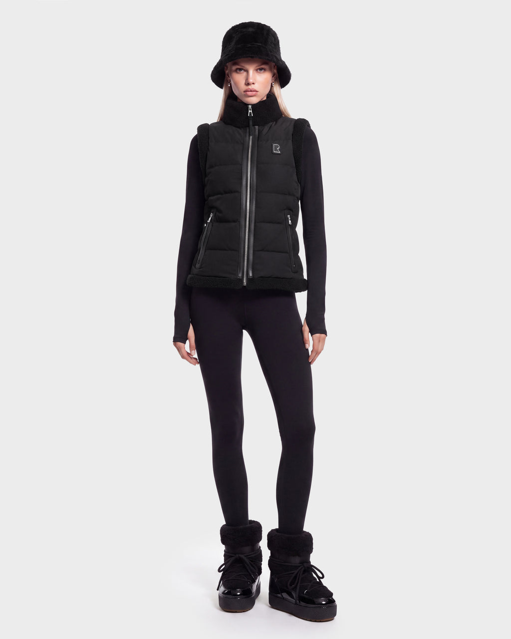 A woman stands facing forward against a plain white background, wearing the Rudsak HALI - 8126771 Black puffer vest filled with RDS-certified down for lightweight warmth, paired with a black bucket hat, long-sleeve black top, black leggings, and black boots.