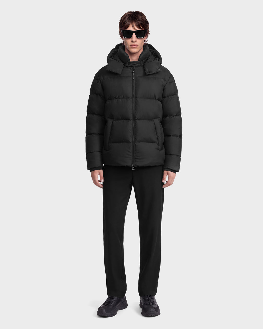 A man wearing a Rudsak JASON - 6126803 Black hooded puffer jacket, black trousers, black sneakers, and sunglasses.