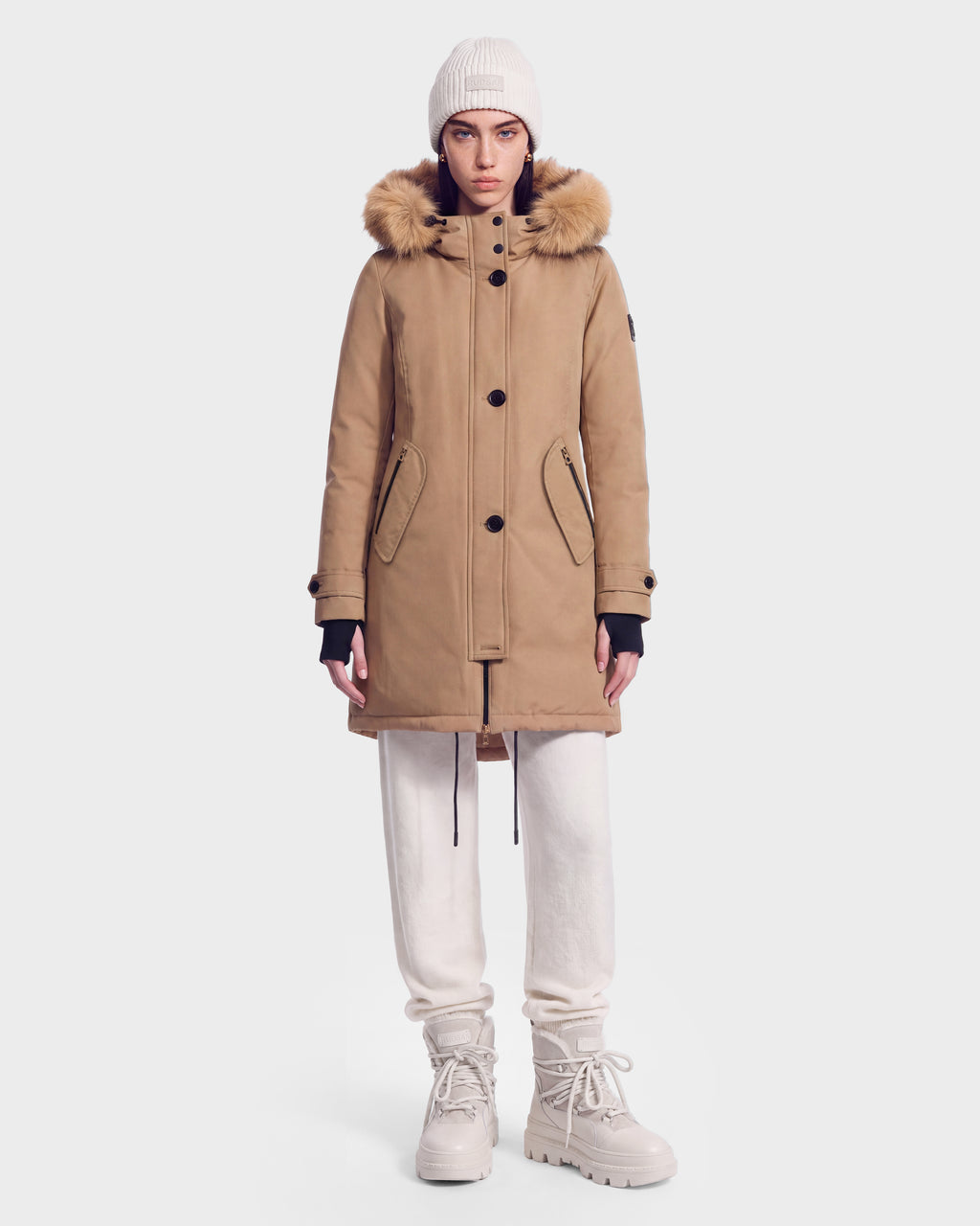 A person wearing the Rudsak KEYSHIA - 8126517 Camel women’s down parka with a fur-lined hood, white knit beanie, white pants, and beige boots stands against a plain white background.
