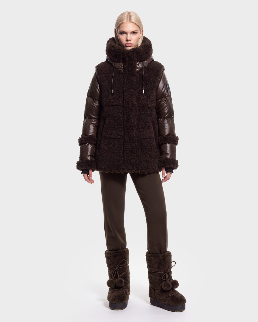 A person with straight blonde hair models the Rudsak MARISSA - 8126728 Espresso, a down-insulated winter coat in a dark, textured finish, styled with matching pants and furry boots with pom-poms, standing against a plain light background.